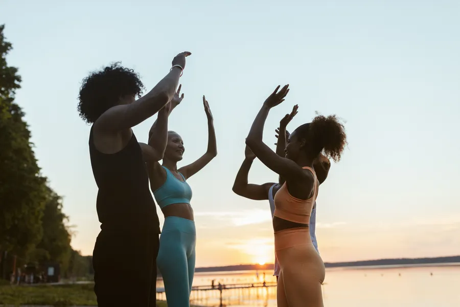 Diverse groep mensen geeft elkaar een high five bij zonsondergang aan het water, symbool voor een gezonde community die samen de regie neemt.