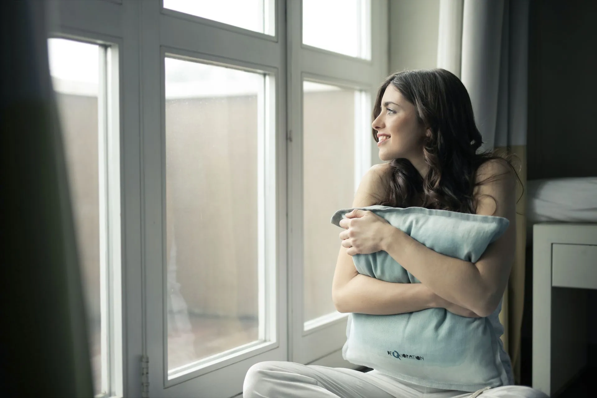 Vrouw zit met een lichtblauw kussen bij het raam en kijkt glimlachend naar buiten