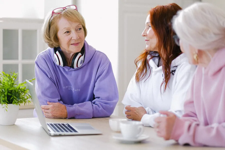 Drie vrouwen praten aan een tafel met een laptop en kopjes