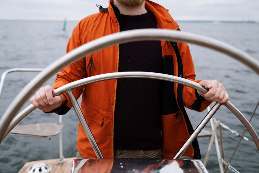 Close-up van een kapitein in oranje jas aan het stuurwiel van een boot op zee.