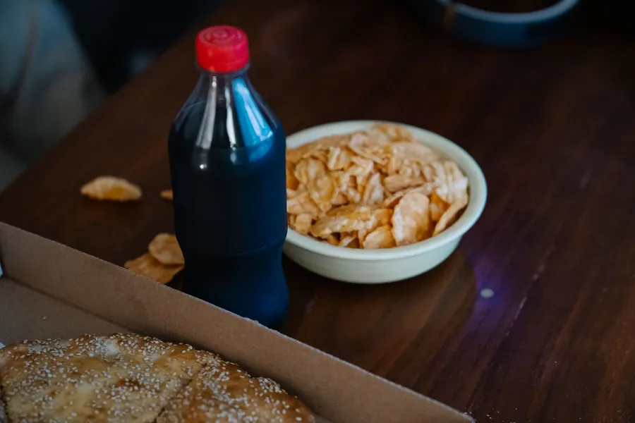Frisdrankfles tussen afhaalpizza en chips, gezien van bovenaf.