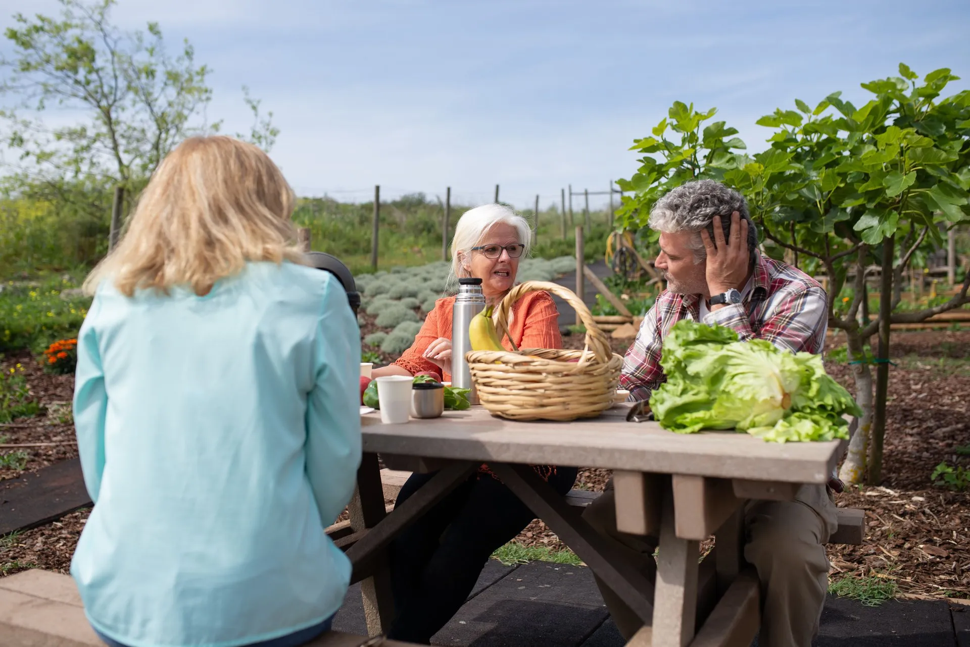 Drie volwassenen zitten met een mand groenten aan een tafel in een tuin