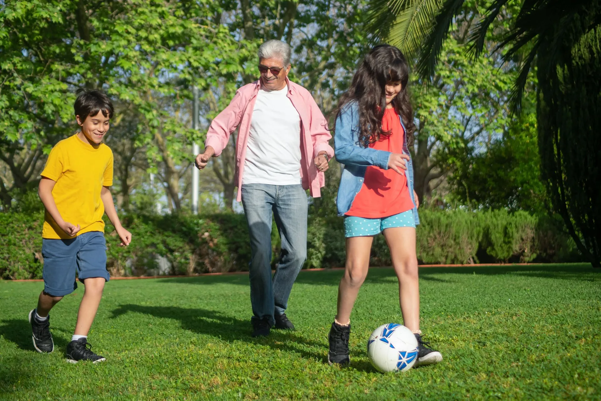 Een oudere man en twee kinderen spelen met een voetbal op gras