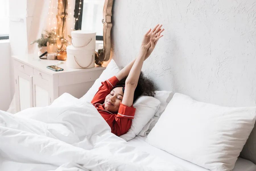 Vrouw in rode pyjama rekt zich uit in een rustige slaapkamer in het ochtendlicht.