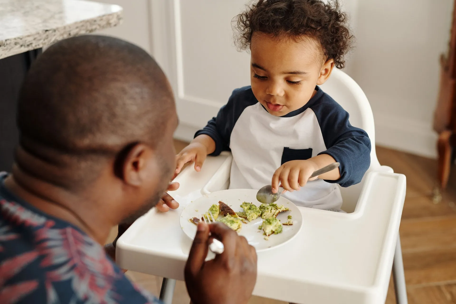 Volwassene zit bij een jong kind dat in een kinderstoel eet
