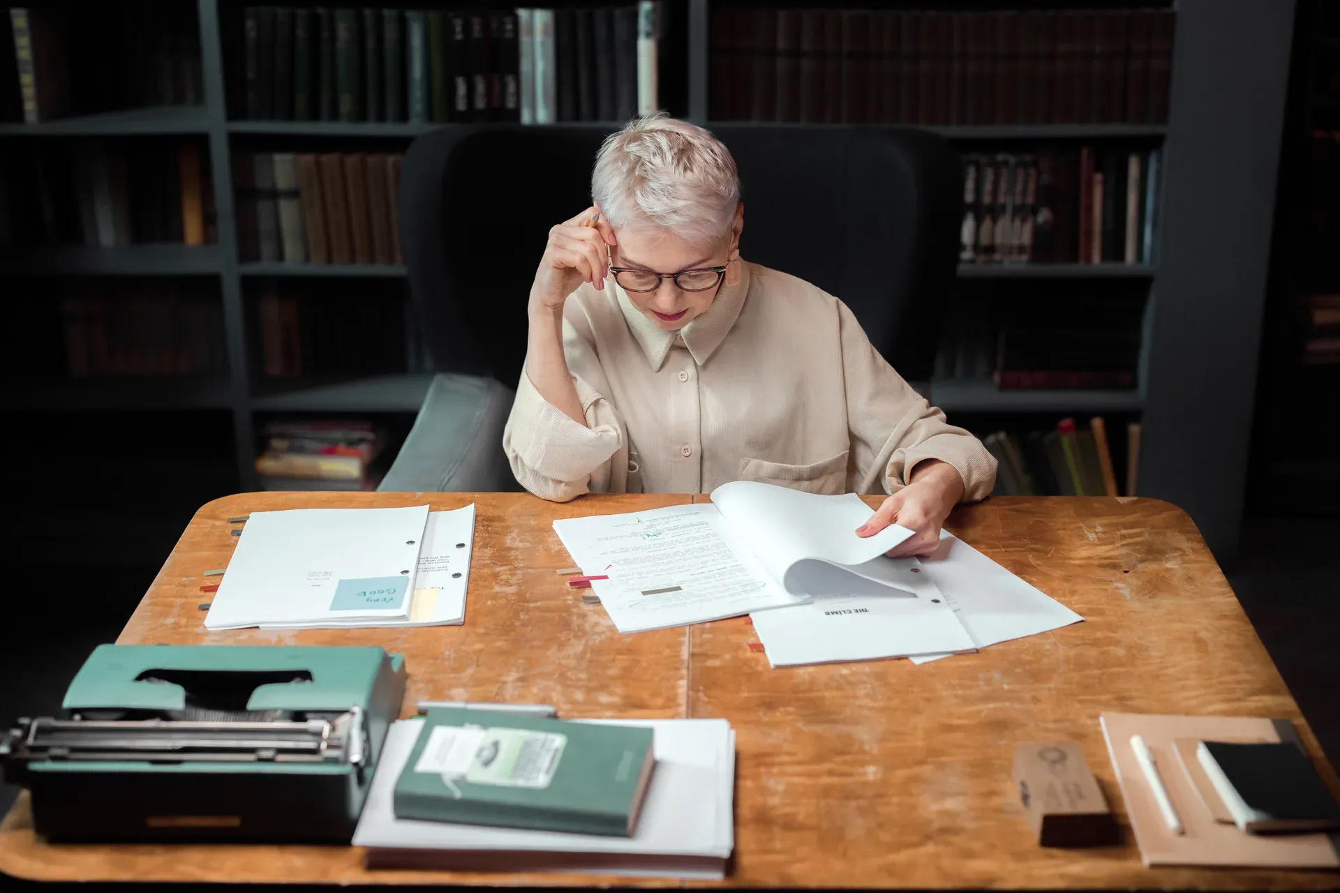 Oudere vrouw met bril leest papieren aan een houten tafel, met een boekenkast op de achtergrond