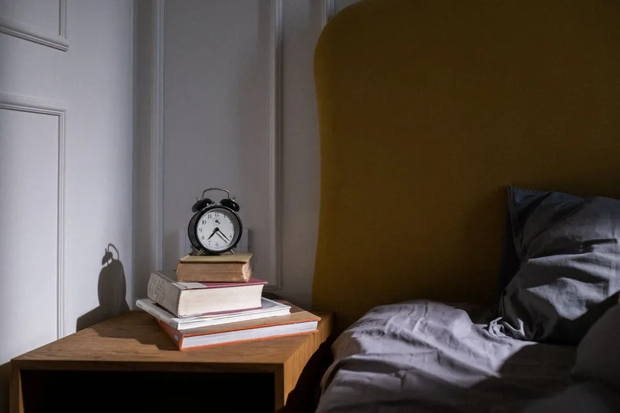 Wekker en boeken naast een bed in een donkere slaapkamer