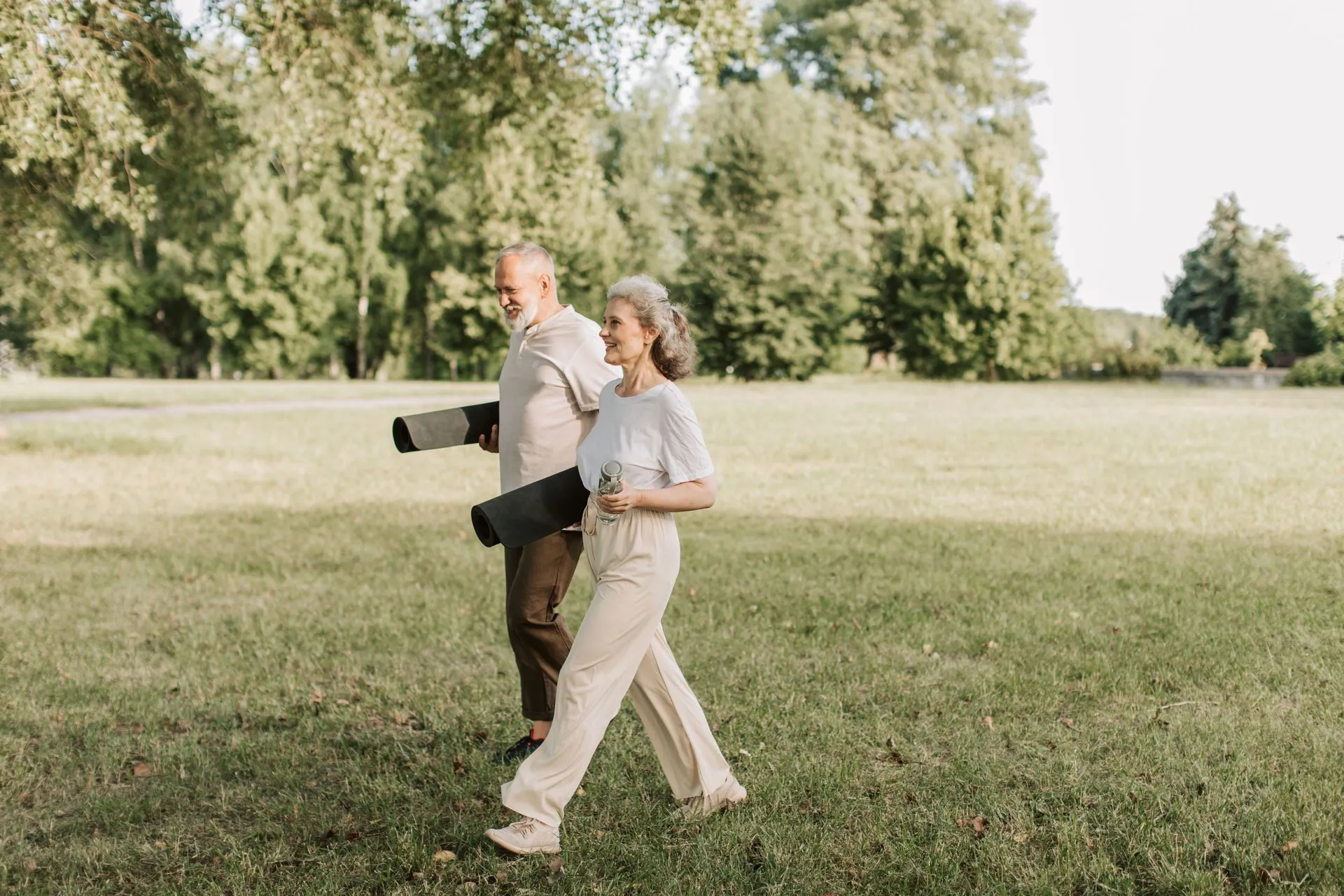 Ouder stel loopt met yogamatten over het gras op een zonnige dag.