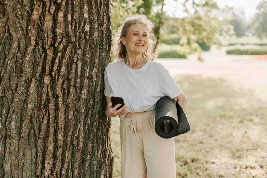 Vrouw met yogamat naast een boom in het park