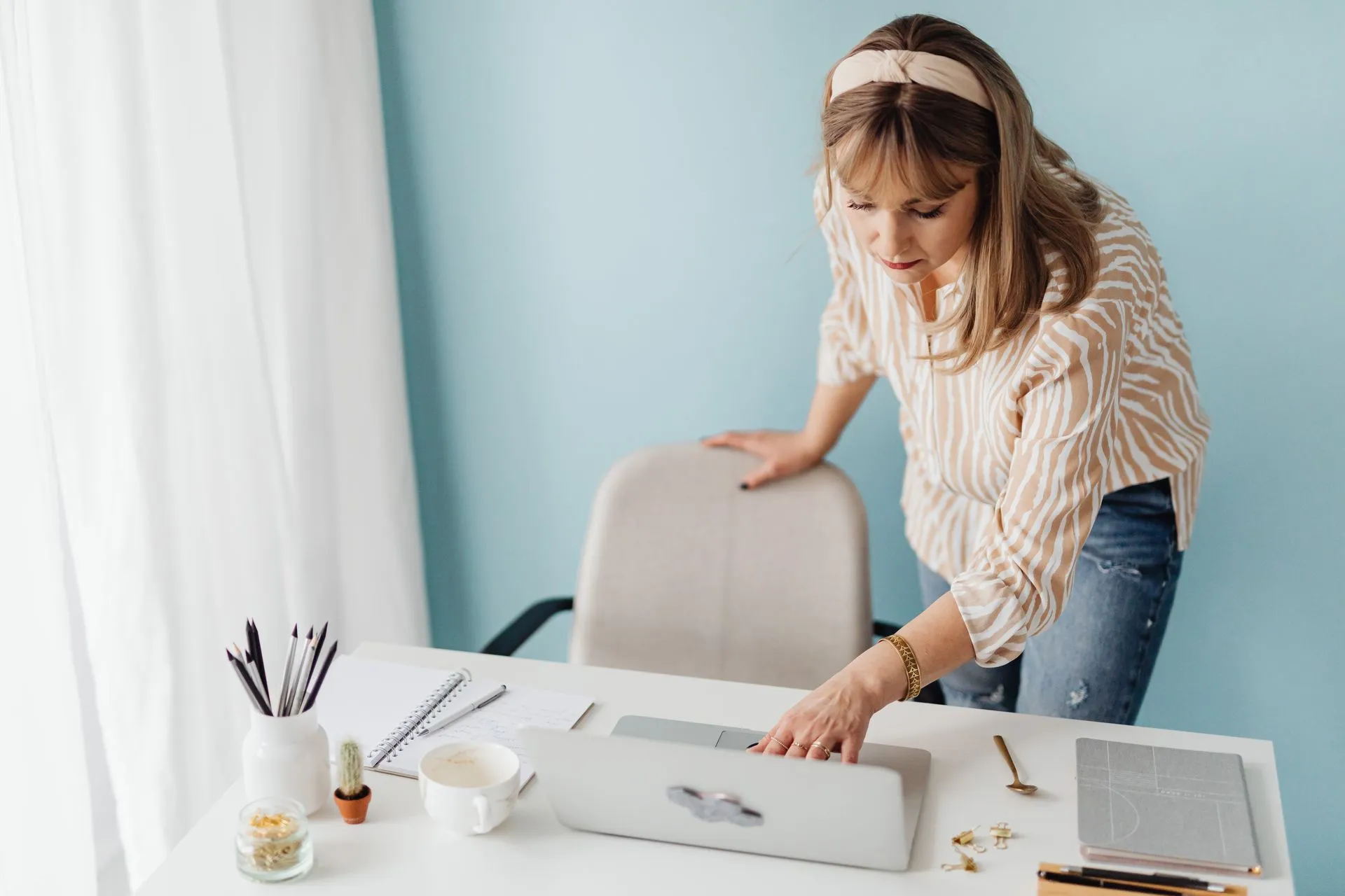 Vrouw werkt staand aan een bureau met laptop, notitieblok en koffie.